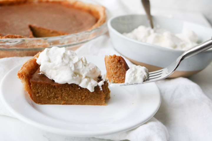 slice of pumpkin pie with whipped cream on top on a white plate with a fork holding a bite 