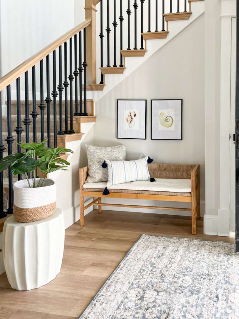 front entry with a bench and a pillow. Seashell art on the walls in black frames with white mats. Cement garden stool holding faux monsterra plant. Blue entry way rug.